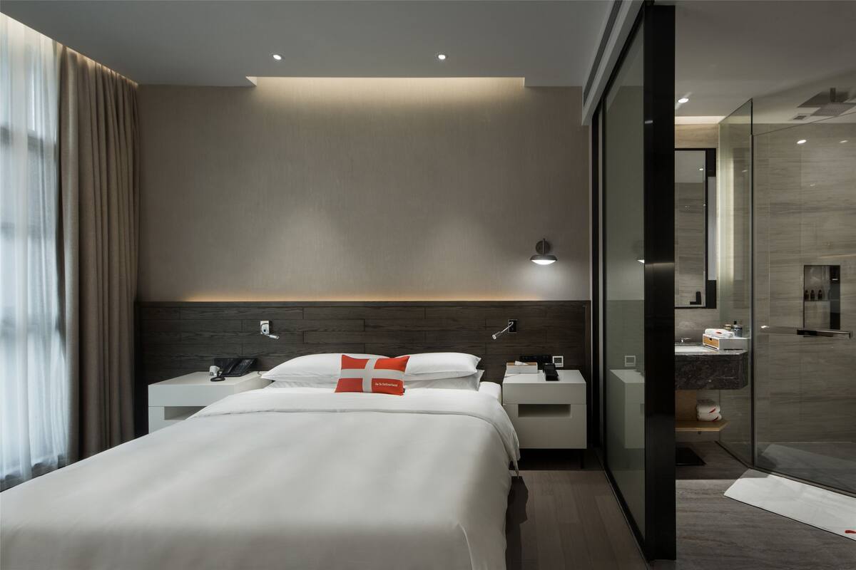Ein modernes Hotelzimmer mit einem großen Bett, Nachttischen, einem Beistelltisch, einer Duschkabine aus Glas und Blick ins Bad.