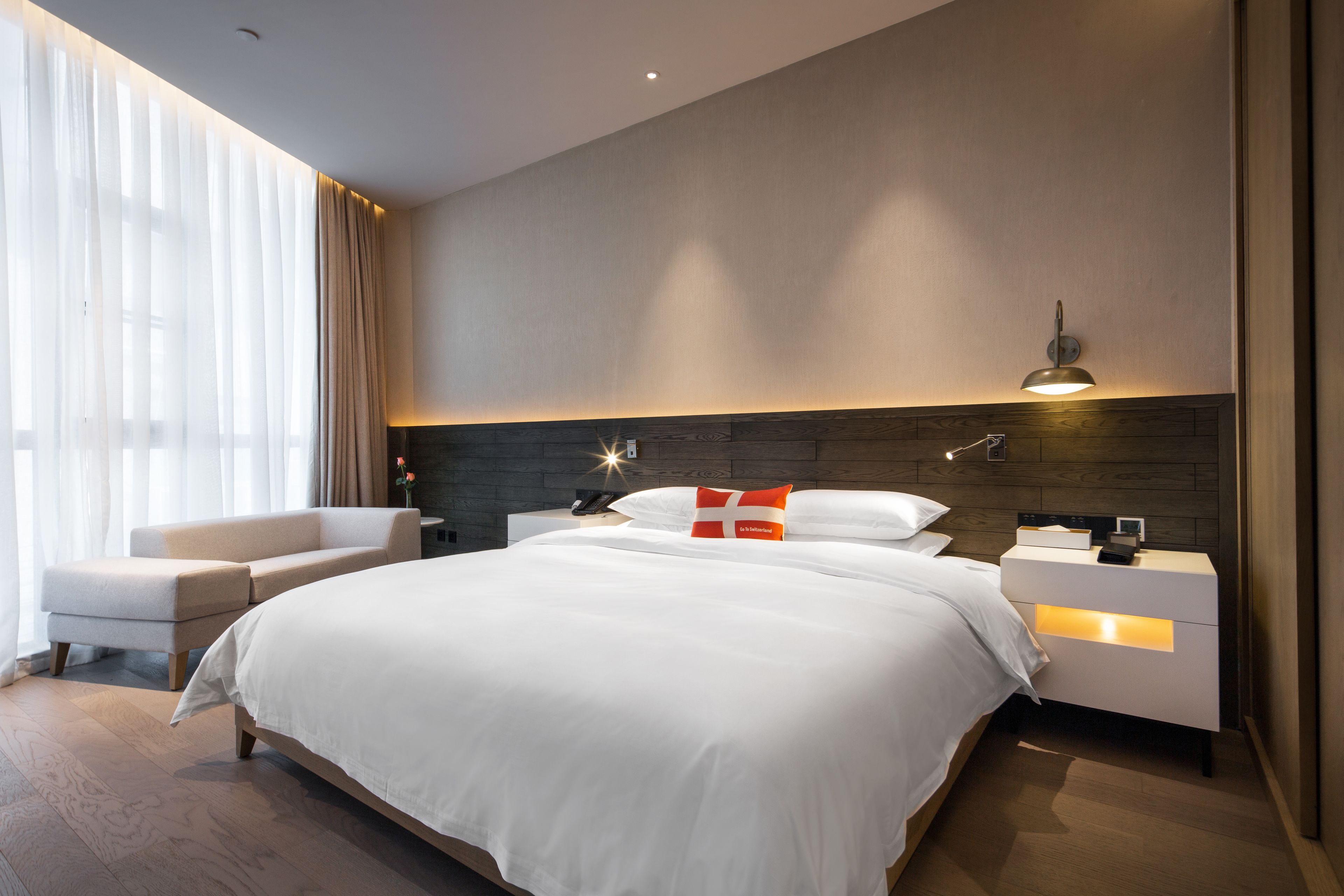Et moderne hotelværelse med en stor seng, et natbord, en sofa og et stort vindue med lette gardiner.