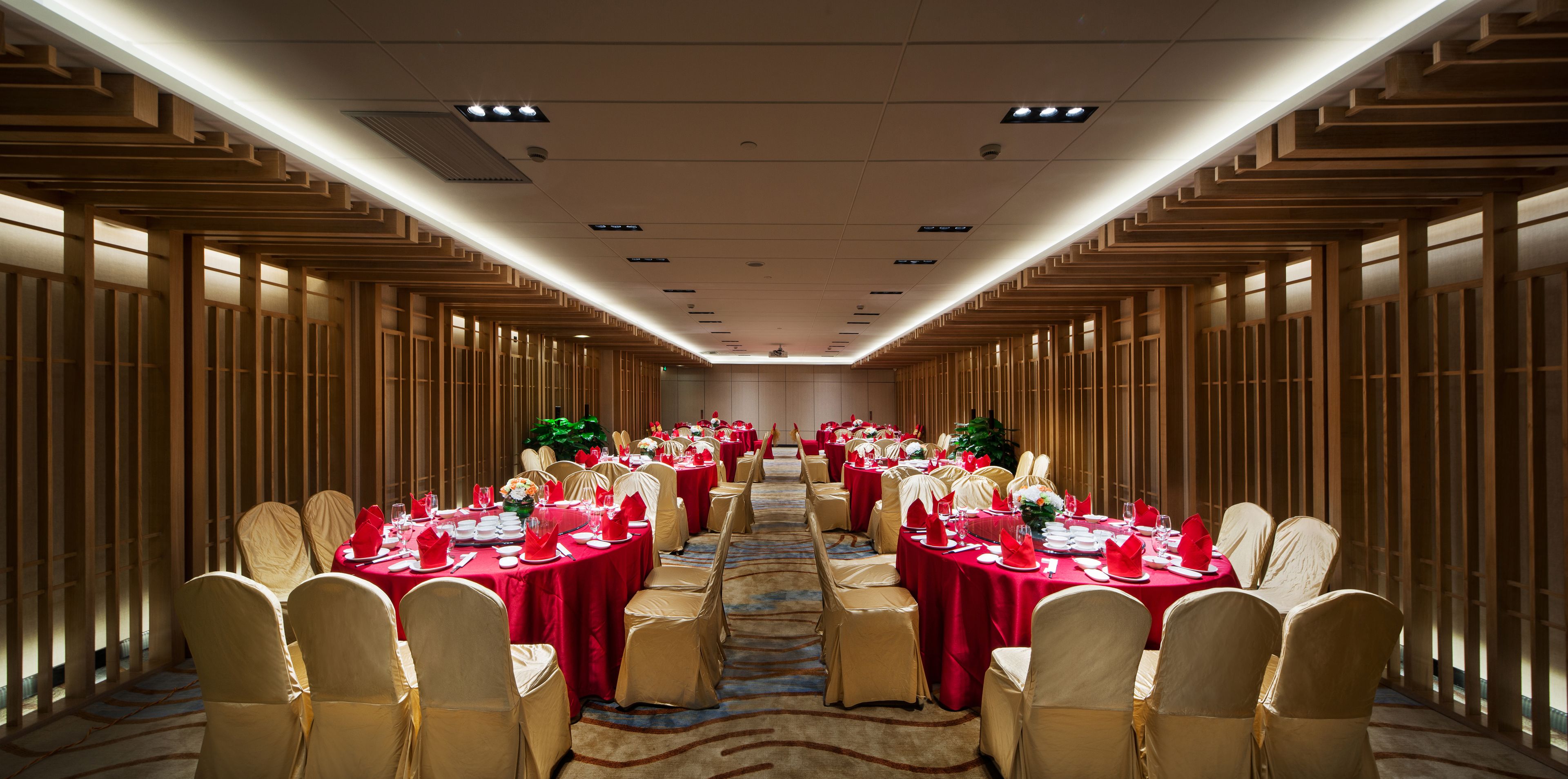 Un salón de banquetes con filas de mesas dispuestas para un evento formal, destacando un pasillo central y paneles de madera en las paredes.