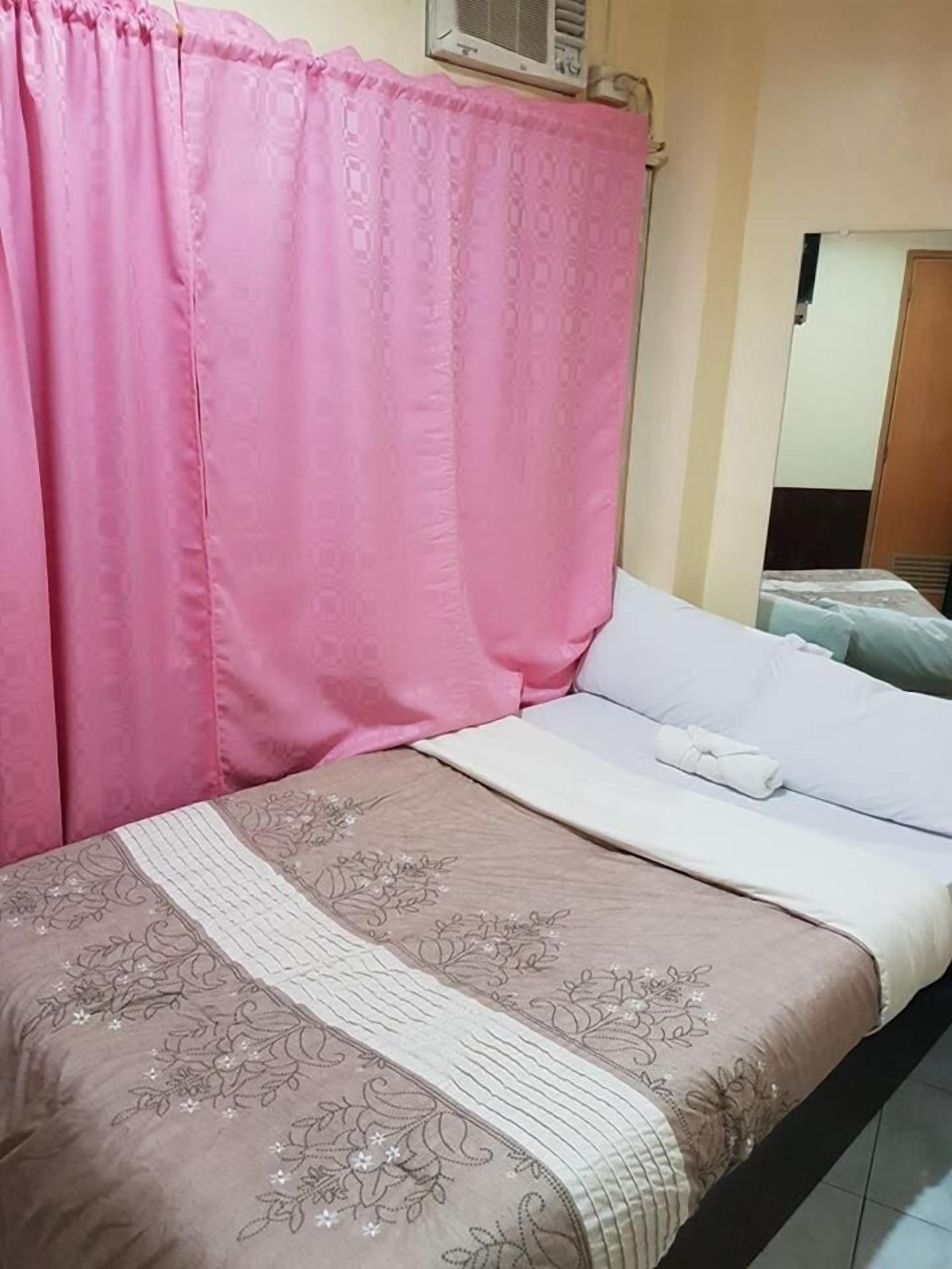 A bed with a floral bedspread, a white pillow, and a pink curtain.