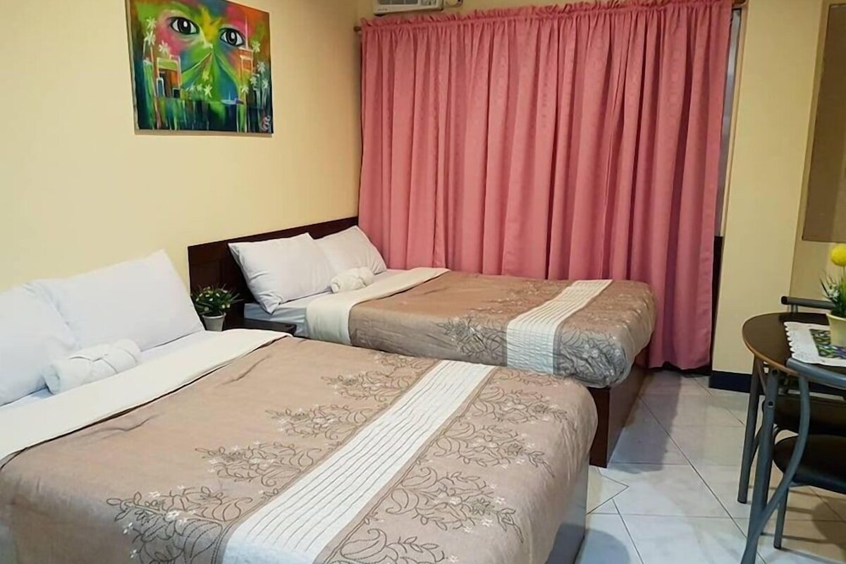 A hotel room with two beds, a painting on the wall, a pink curtain, and a small table with a chair.