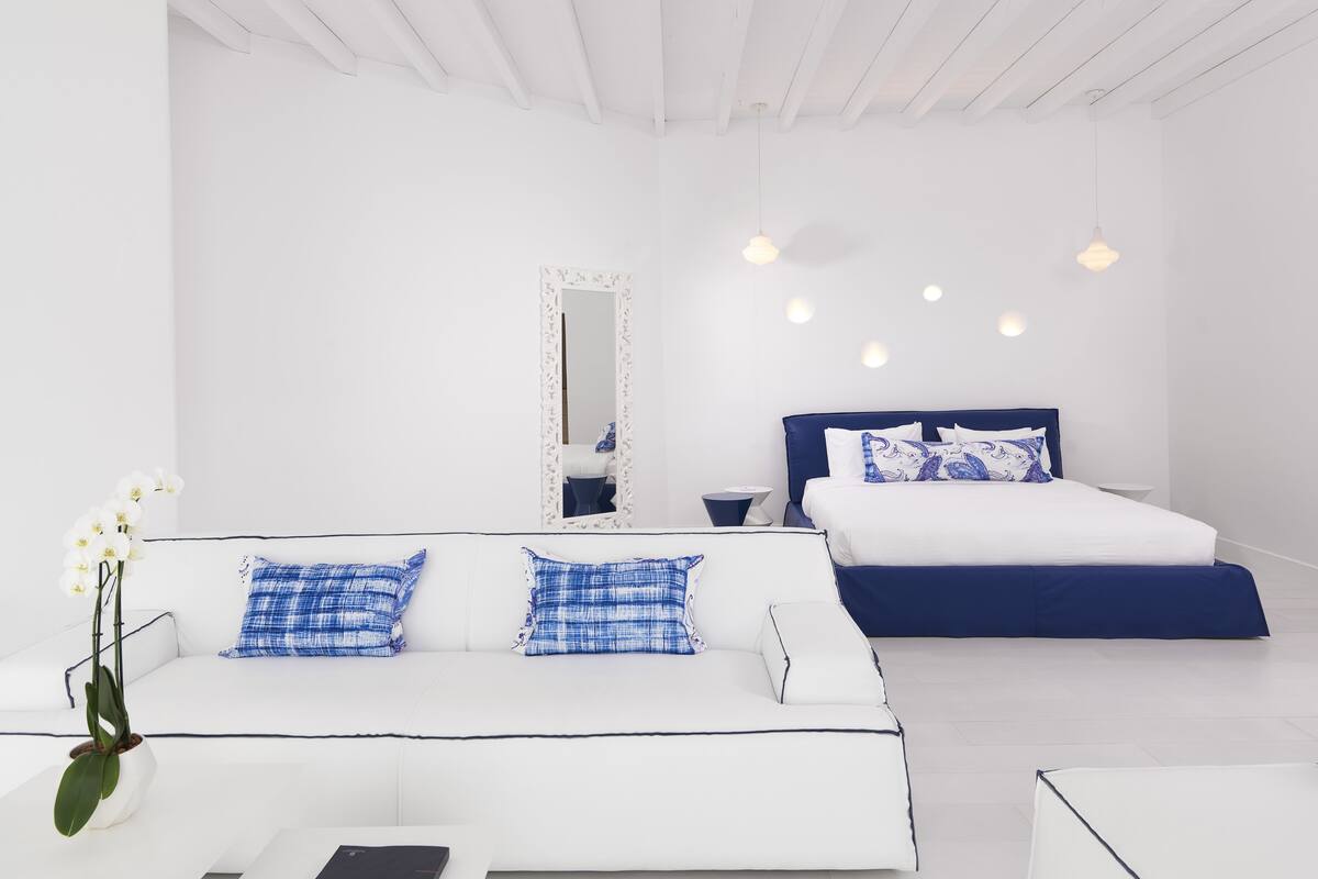 Ein modernes Schlafzimmer mit weißer Decke, einem großen Bett mit blauer Bettwäsche, einem weißen Sofa mit blauen Kissen und einem weißen Boden.