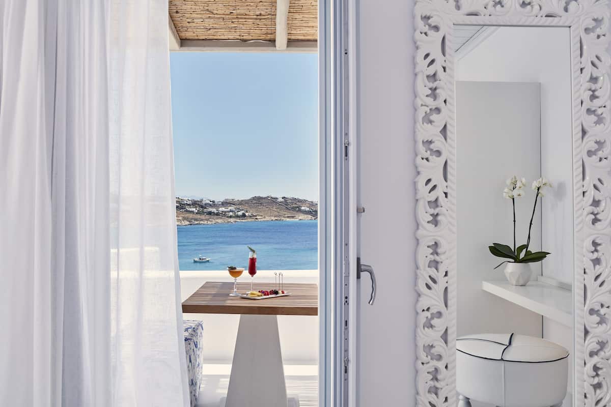 Ein Zimmer mit Meerblick, ein Holztisch mit Getränken und Obst, sowie ein weißes Badezimmer mit Spiegel und Orchidee.