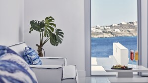 A modern living room with a white sofa, a potted plant, and a dining table with a plate of food and two drinks. A large window offers a view of the sea and buildings. - Katikies Mykonos - The Leading Hotels Of The World (Mykonos)
