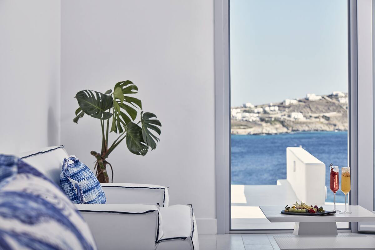 Ein modernes Wohnzimmer mit einem weißen Sofa, einer Topfpflanze und einem Esstisch mit einer Speiseplatte und zwei Getränken. Ein großes Fenster bietet Blick auf das Meer und Gebäude.