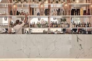 Uma área de bar com bancadas de mármore, prateleiras cheias de garrafas de bebidas e luminárias pendentes de cobre.