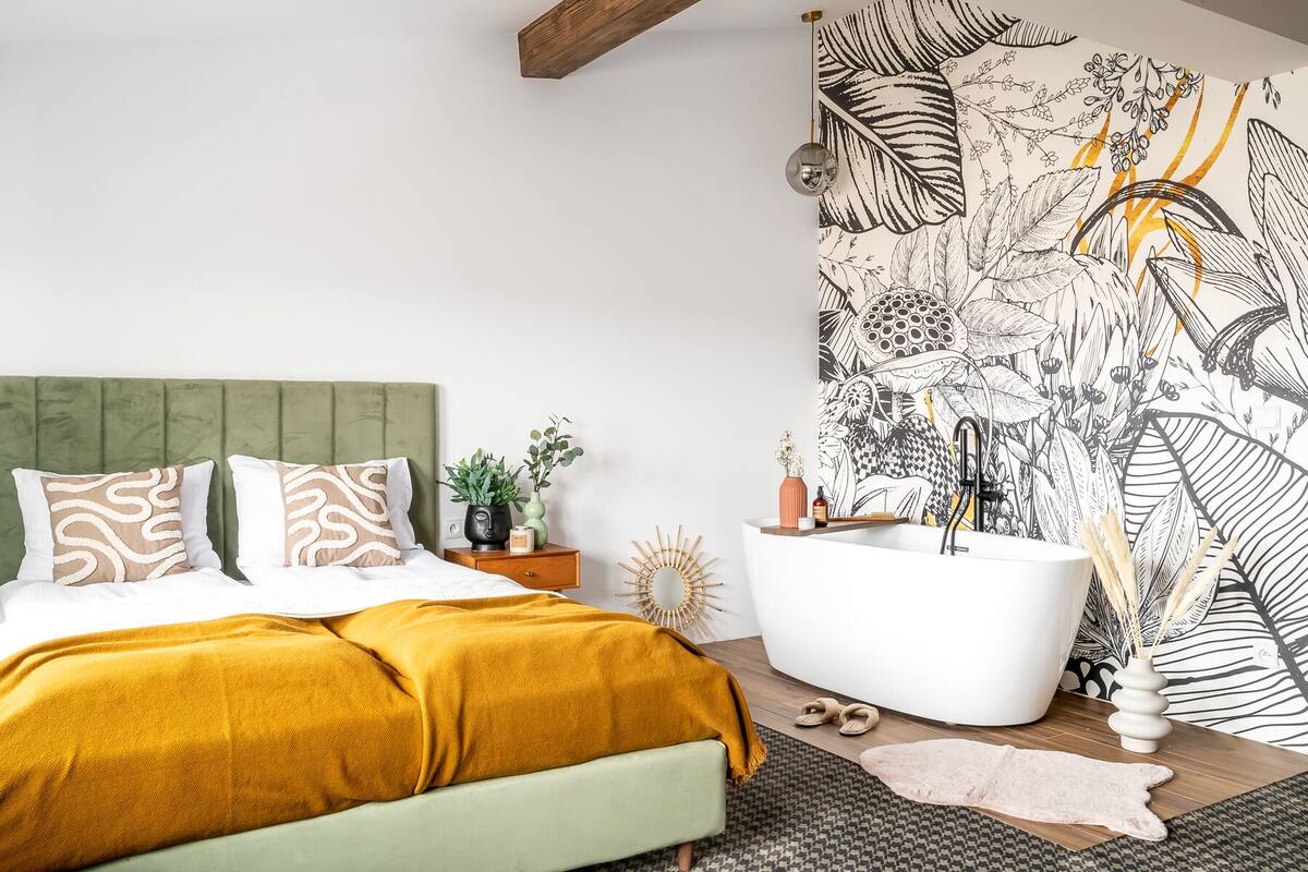 A modern bedroom with a green upholstered headboard, a large bed with a mustard-colored blanket, a white bathtub, and a wall mural with botanical illustrations.
