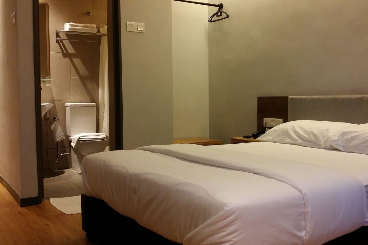 A hotel room with a bed, a bedside table with a phone, a nightstand, a towel rack, and a shower area.