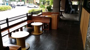 An outdoor seating area with wooden benches and round tables, a tiled floor, and a view of the street with parked cars and greenery.