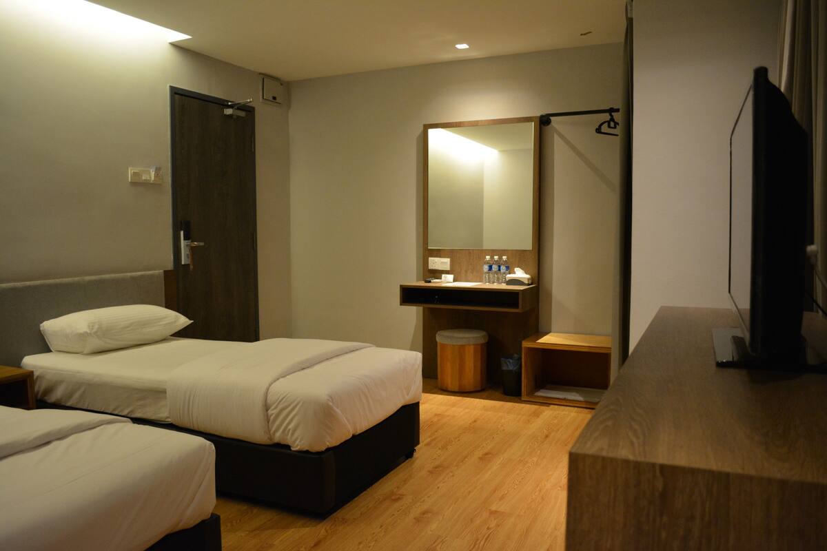 A hotel room with two beds, a wooden floor, a large mirror, a sink, and a television.