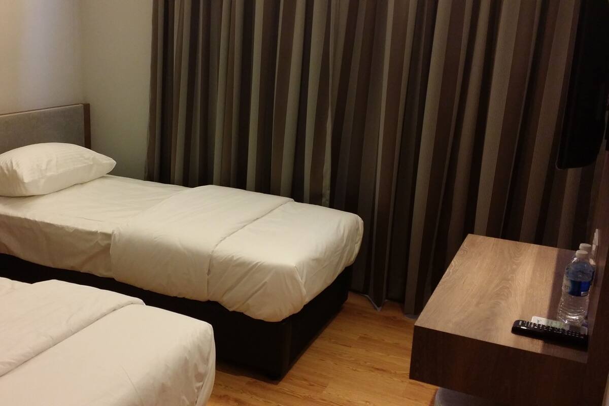 A hotel room with two single beds, a wooden nightstand with a phone, a wooden table with a remote control, and a water bottle.