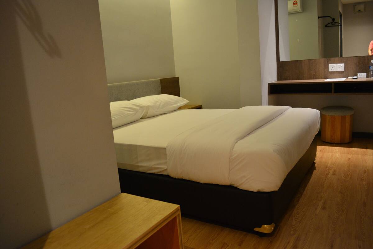A hotel room with a bed, a wooden nightstand, a small stool, a mirror, and a wooden floor.