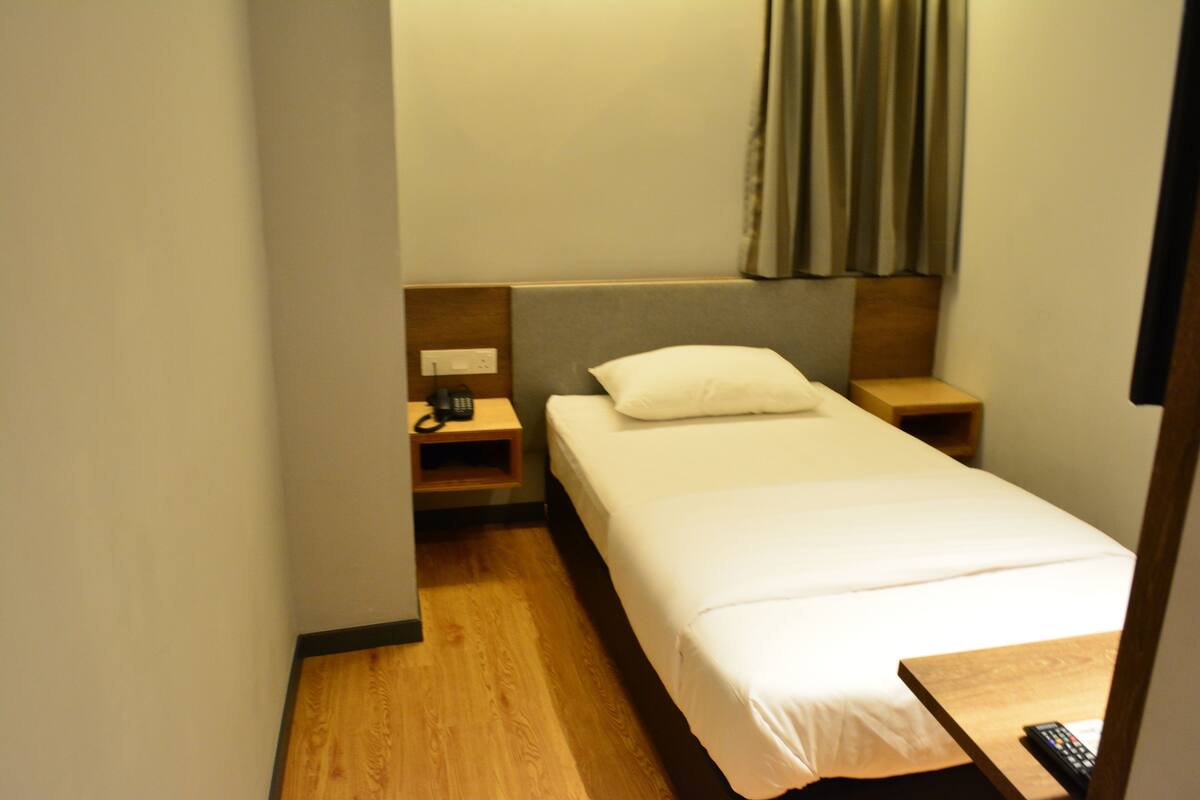 A small hotel room with a single bed, a nightstand, a bedside table, and a remote control on the table.