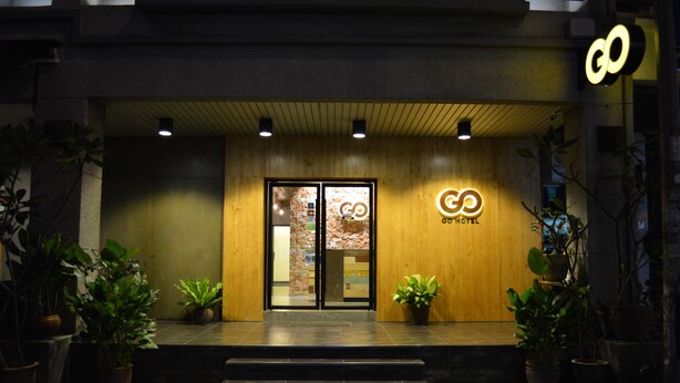 A hotel entrance with a wooden facade, glass doors, and a sign that reads "GO HOTEL". There are potted plants on either side of the entrance.