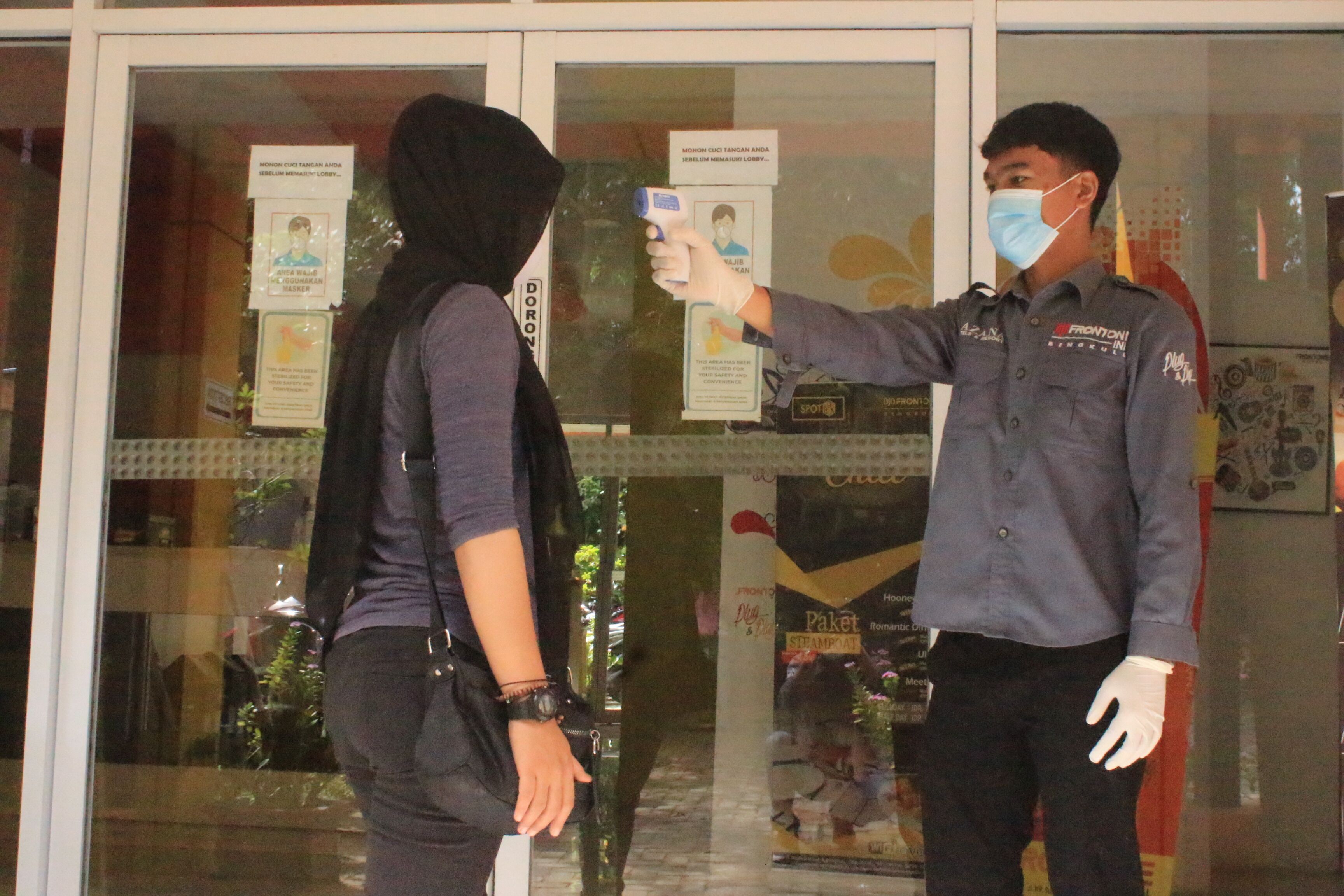 Une personne portant un masque et un uniforme se tient devant une porte vitrée ornée d’affiches.