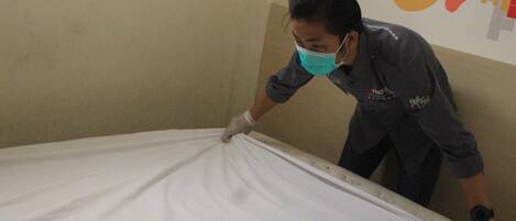 Una persona arreglando una cama en una habitación con una pared lisa y un cartel colorido.