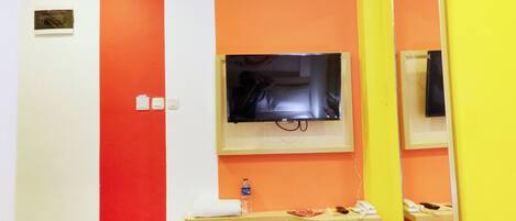 A hotel room with a bed, a TV mounted on the wall, a small shelf with toiletries, and a mirror reflecting part of the room.