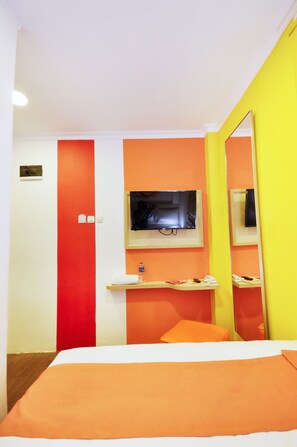 A hotel room with a bed, a TV mounted on the wall, a small shelf with toiletries, and a mirror reflecting part of the room.