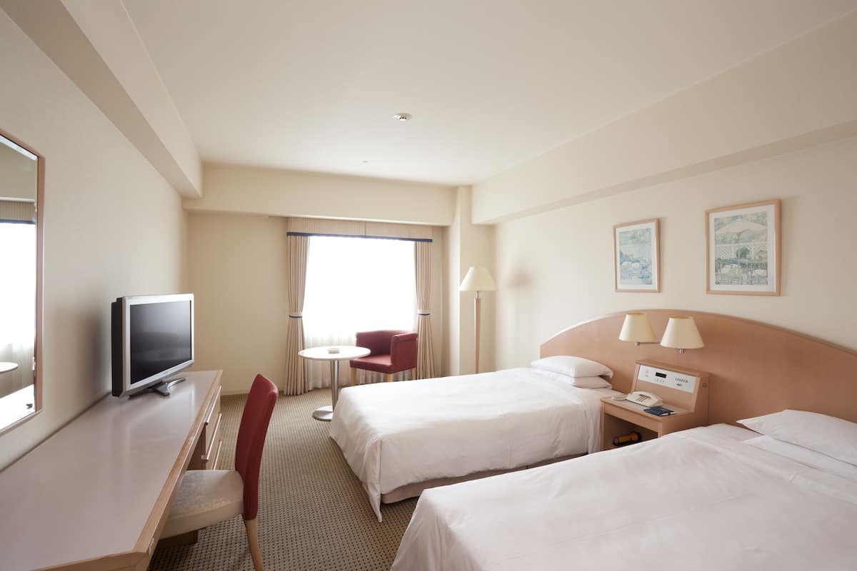 A hotel room with two beds, a desk with a TV, a chair, a small table, and a window with curtains.