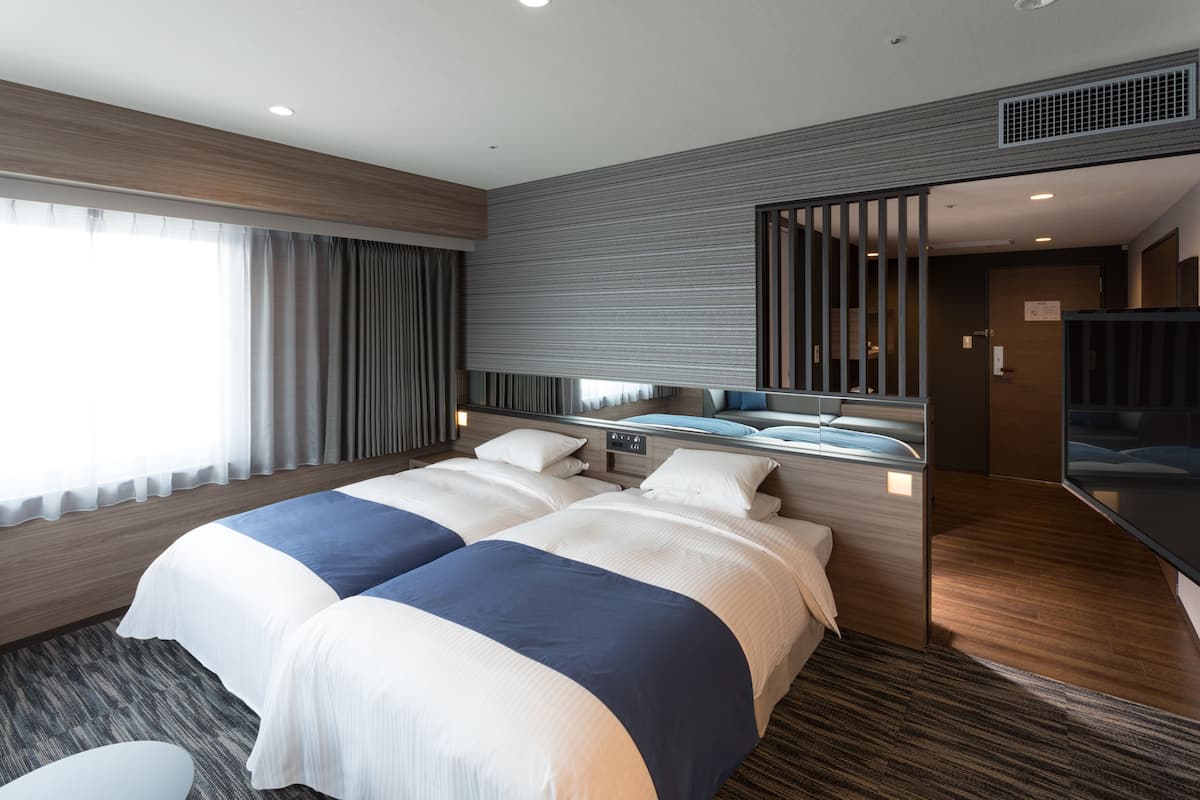 A modern hotel room with two beds, a large window with curtains, a flat-screen TV, and a wooden floor.