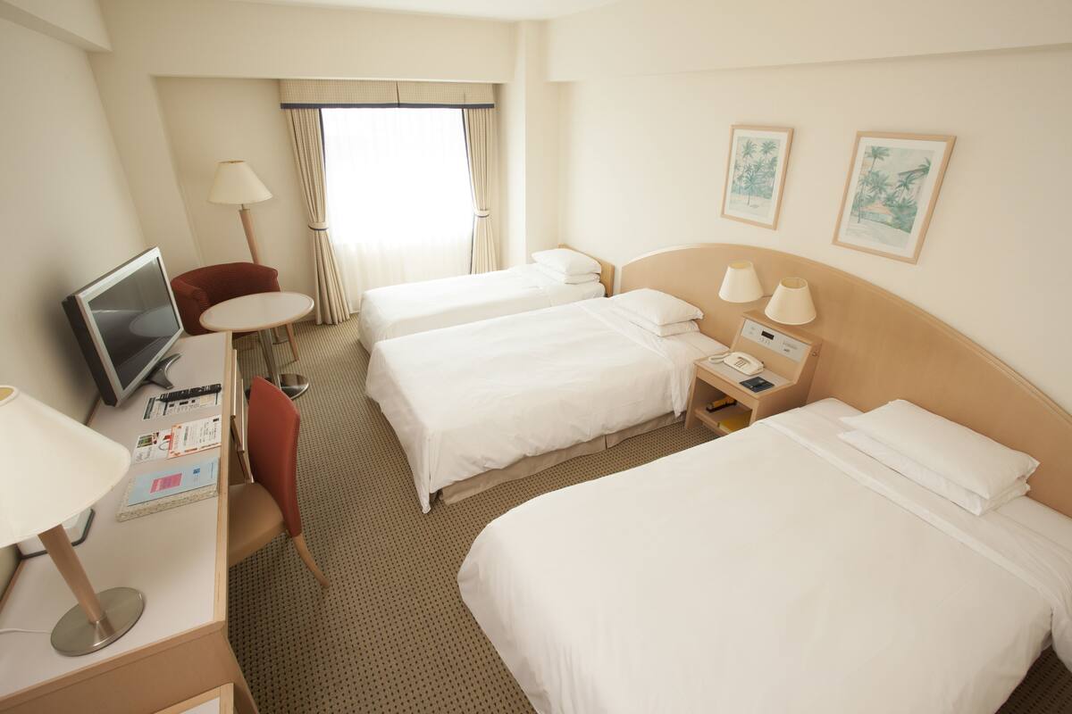 A hotel room with two beds, a desk with a computer, a chair, a lamp, and a window with curtains.