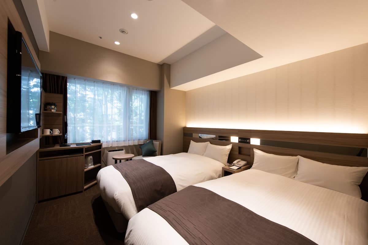 A hotel room with two beds, a desk, a chair, a TV, and a window with curtains.