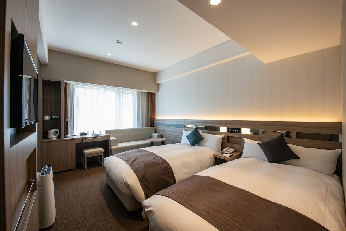 A modern hotel room with two beds, a desk, a TV, and a window with curtains.