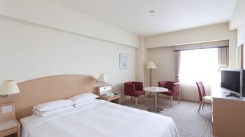 Quarto de hotel com cama, escrivaninha, televisão e duas cadeiras.