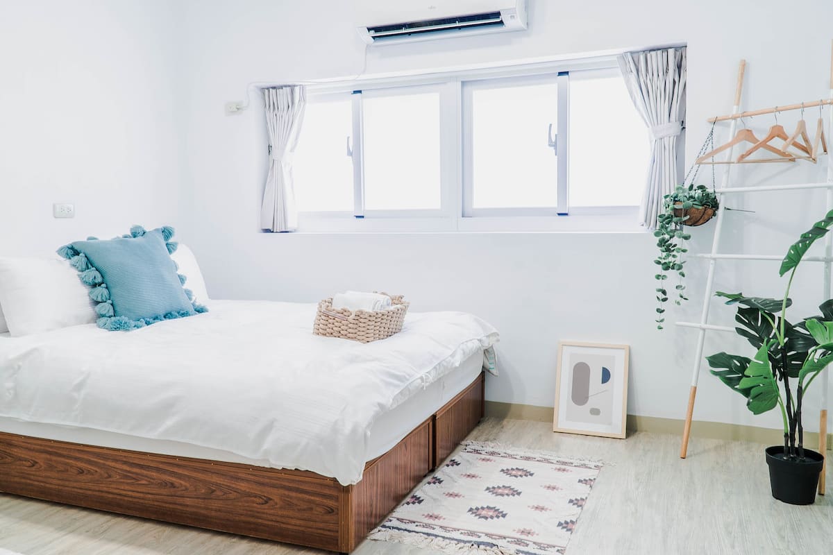 A bedroom with a wooden bed, white bedding, a window with curtains, a hanging plant, a framed picture, and a small rug.