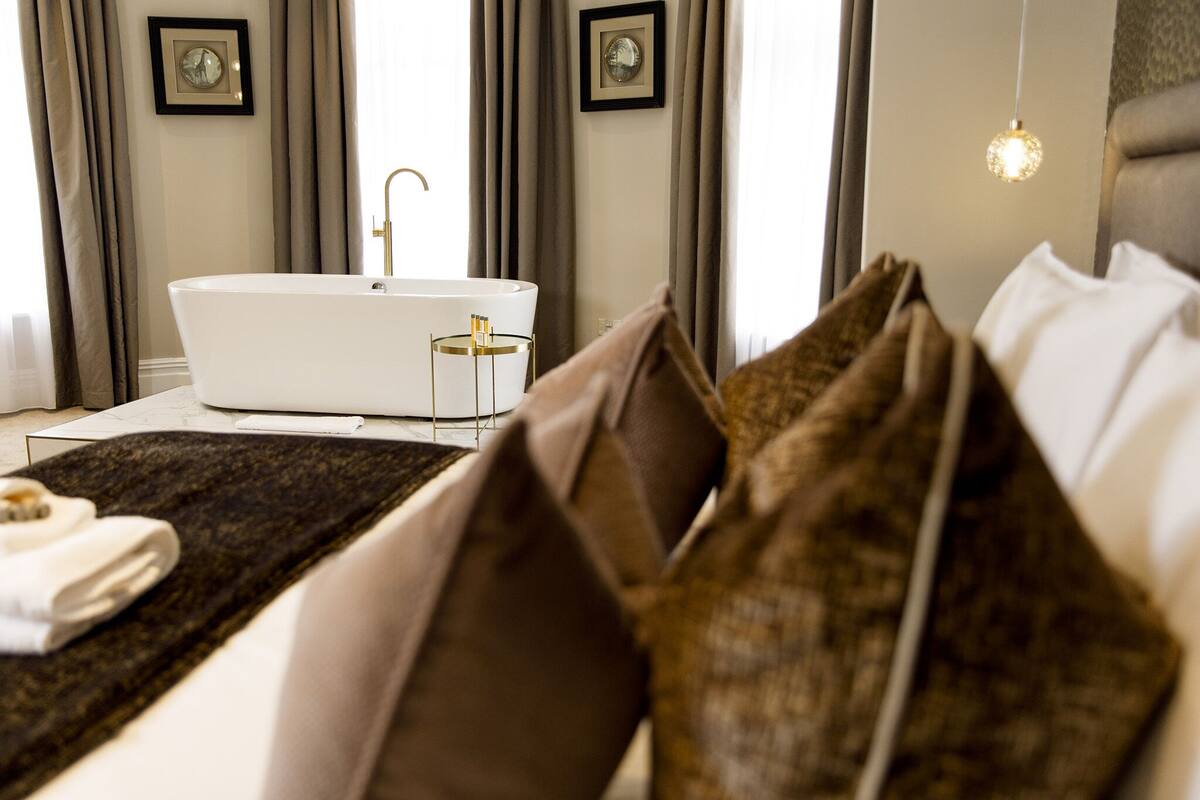 A modern hotel room with a large bed, a white bathtub, and a bedside table with a lamp.
