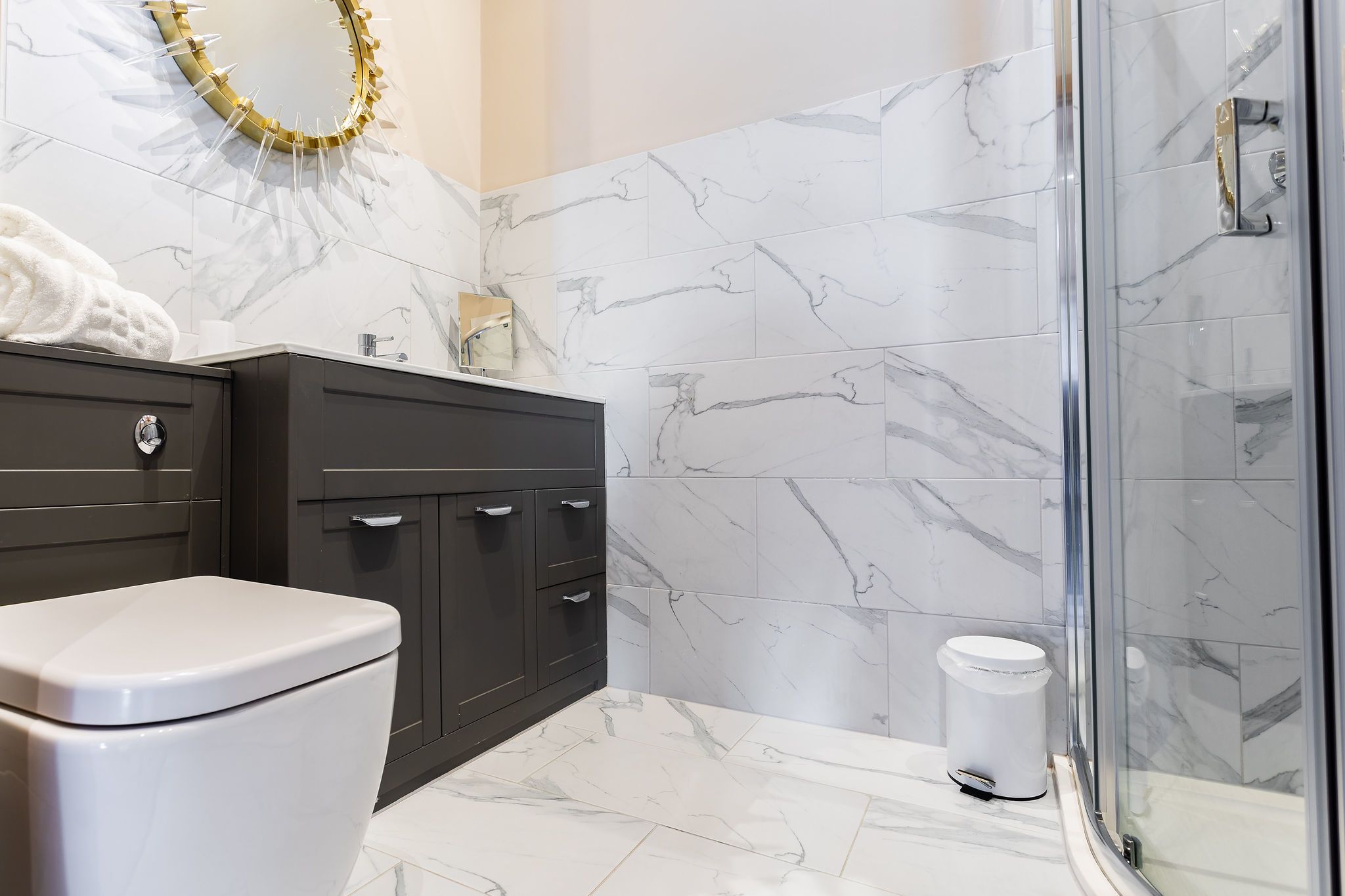 A bathroom with a toilet, dark wooden cabinet, marble tiles, and a glass shower enclosure.