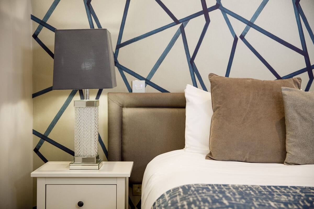 A hotel room with a bed, a nightstand, a lamp, and a geometric-patterned wall.