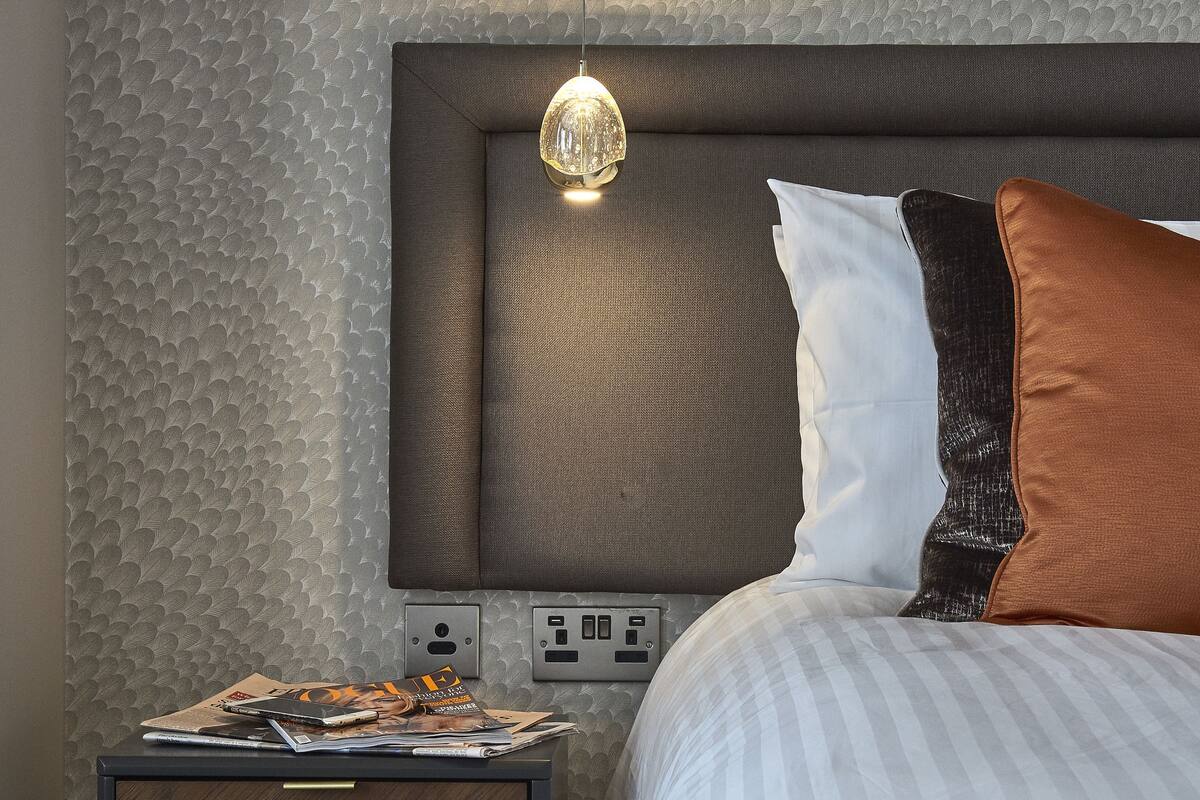 A neatly made bed with a headboard, a bedside table with a lamp, and a power outlet.