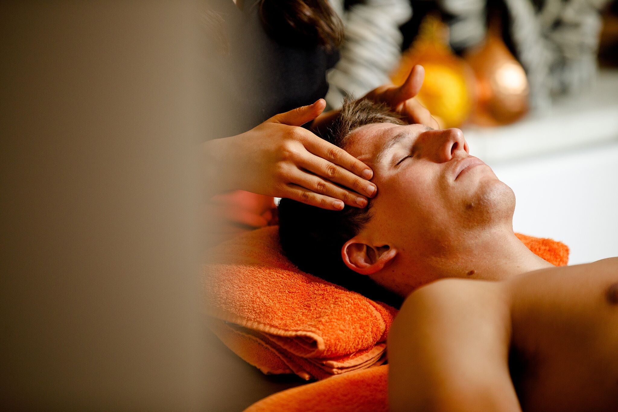 A person lying on a bed with an orange towel, receiving a head massage.