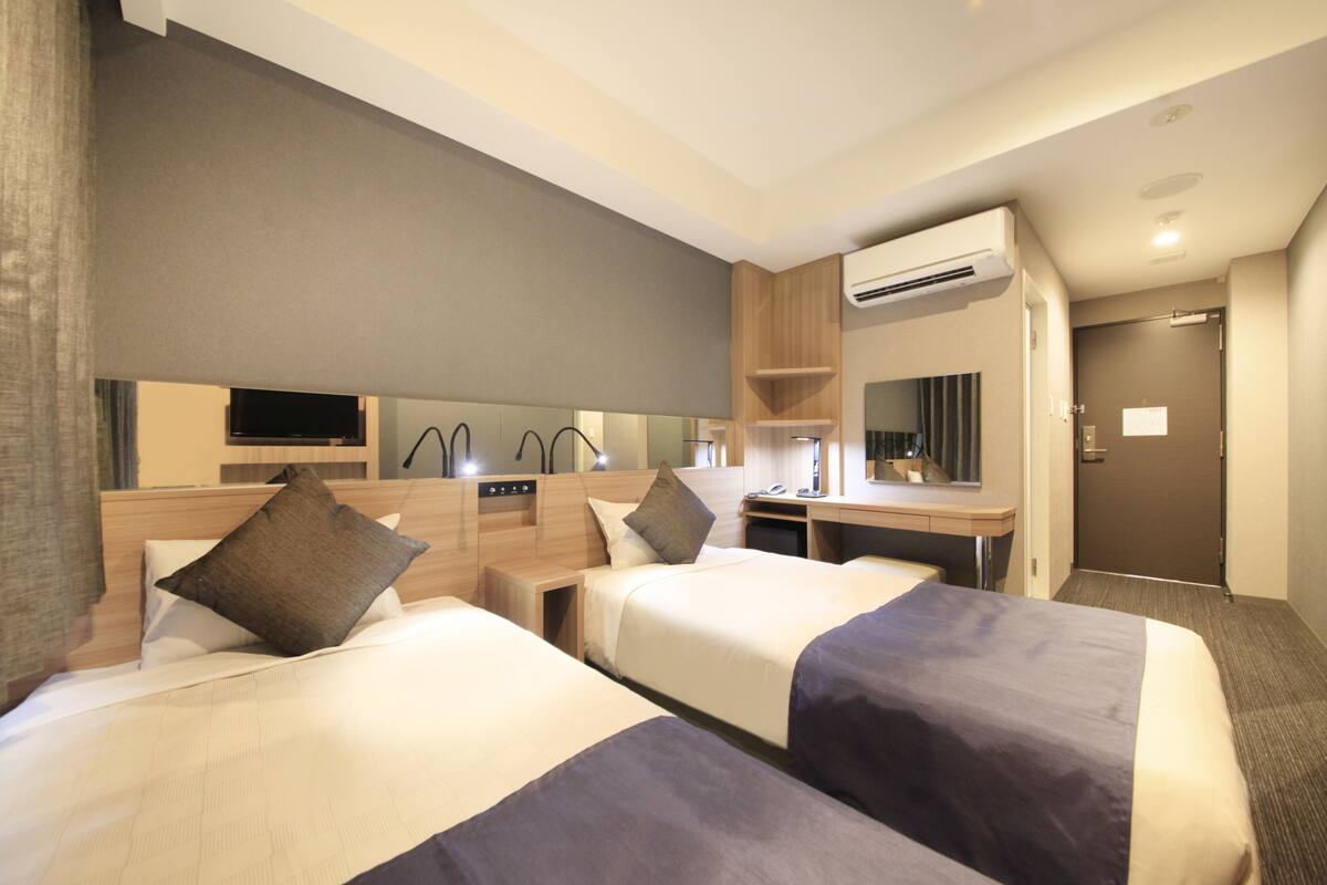 A hotel room with two beds, a desk, a mirror, and an air conditioning unit.