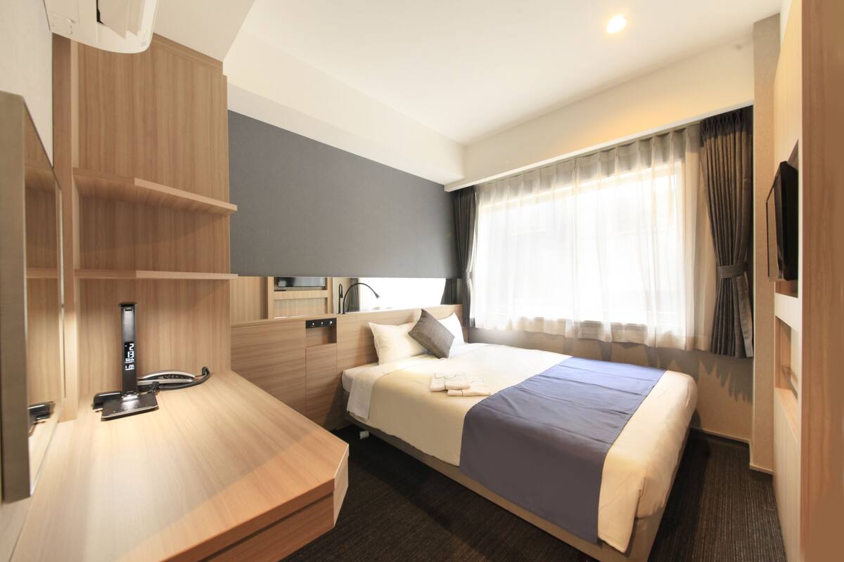 A hotel room with a bed, a desk, a mirror, a TV, and a window with curtains.