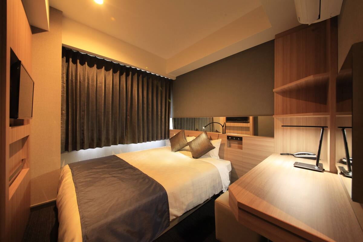A hotel room with a bed, bedside table, desk, and a large window with curtains.