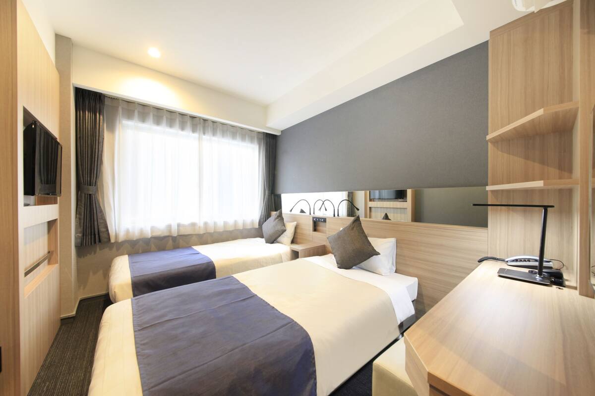 A hotel room with two beds, a desk, a TV, and a large window with curtains.