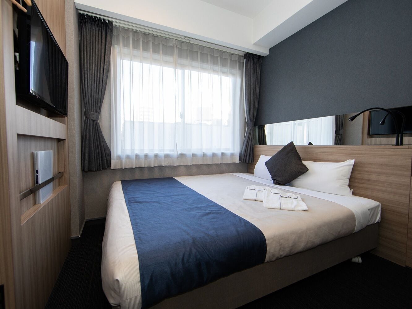 A hotel room with a bed, a TV, a desk with a computer, and a window with curtains.