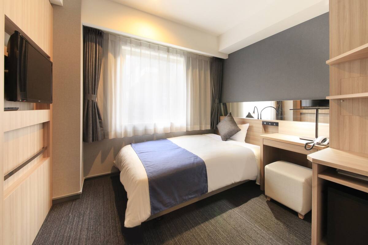 A modern hotel room with a large bed, a desk, a chair, a TV, and a window with curtains.