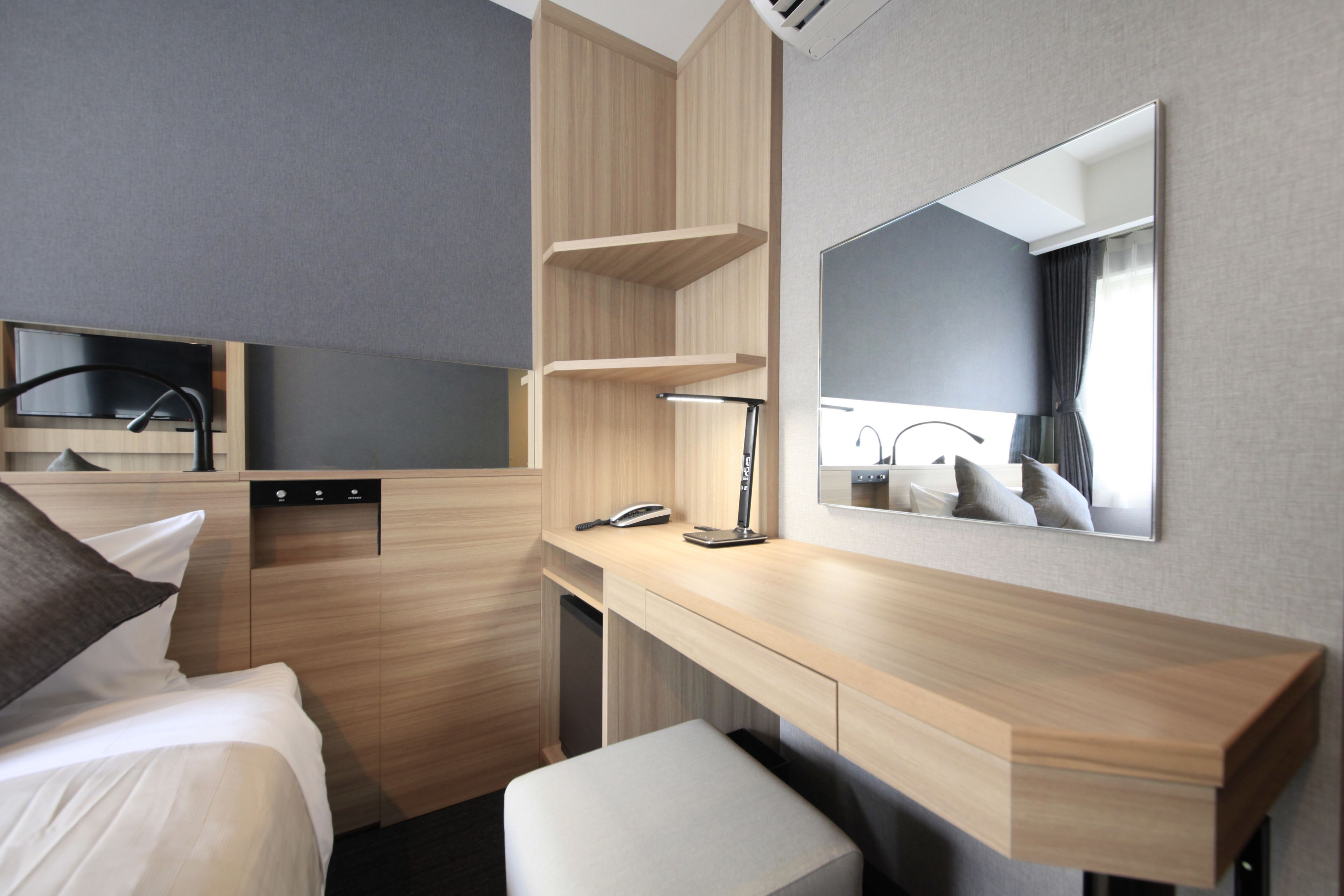 A modern hotel room with a bed, a desk, a mirror, and built-in shelves.
