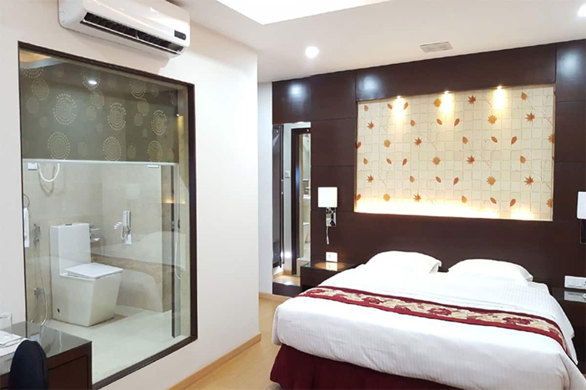 A hotel room with a glass-enclosed shower, a bed with white bedding, a decorative wall panel, and a bedside table with a lamp.