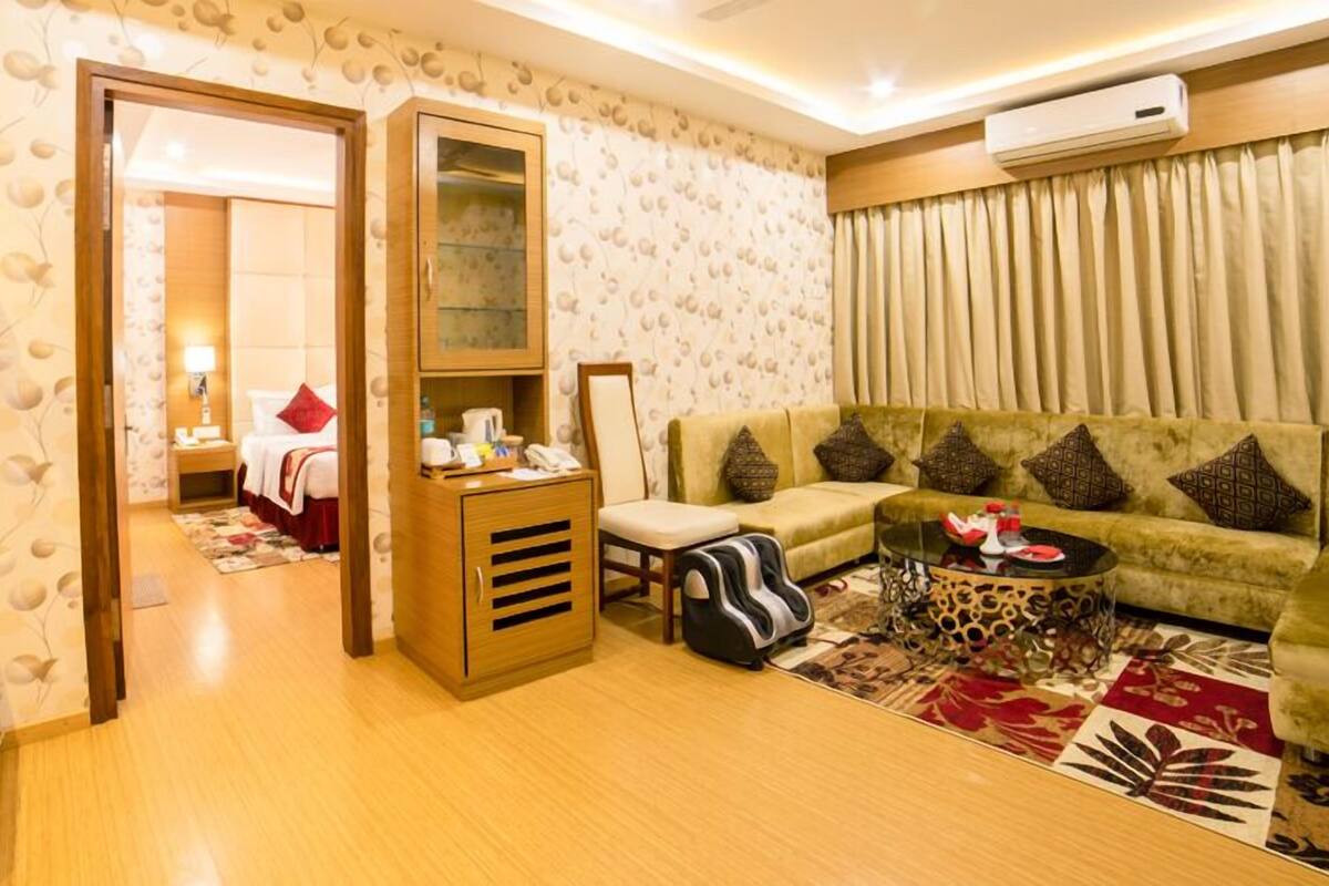 A hotel room with a sofa, a small table, a cabinet, a chair, and a bed in the background.