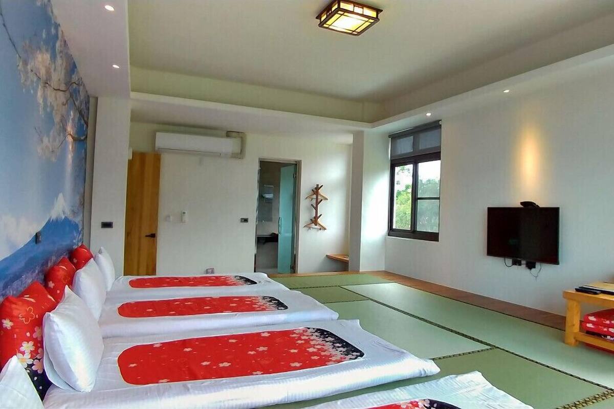 A room with tatami mats, a futon bed with red and white bedding, a TV on the wall, and a large painting on the wall.