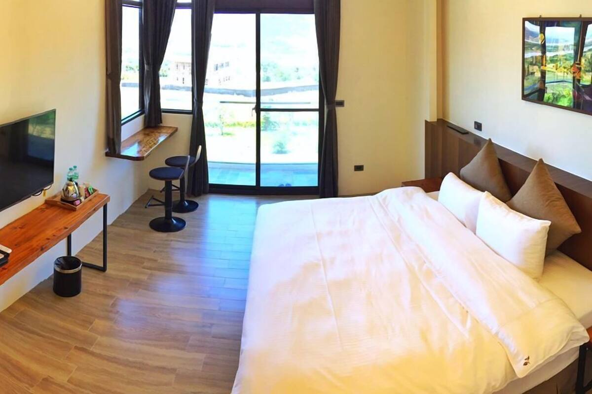 A hotel room with a large bed, a TV, a desk with a chair, and a view of the outdoors through the window.