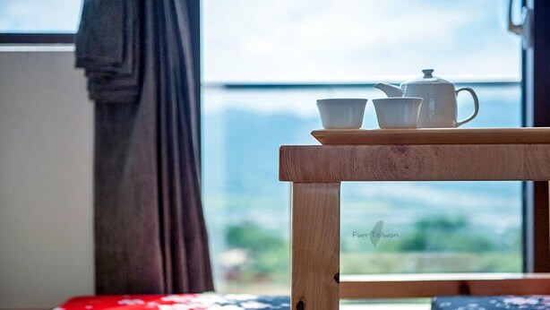 Una mesa de madera con un calentador de agua, dos tazas y un plato, cerca de una ventana con vistas a la vegetación y montañas.