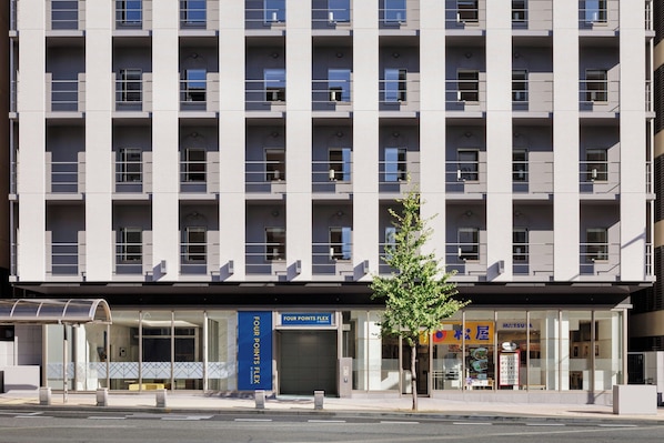 Ein modernes, mehrstöckiges Gebäude mit Glasfassade, einem blauen Schild mit der Aufschrift 'Four Points Flex' und einem Baum davor.