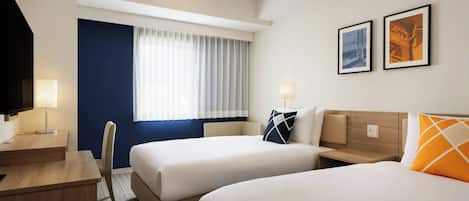Habitación de hotel con dos camas, un escritorio, una silla, un televisor y dos cuadros en la pared.
