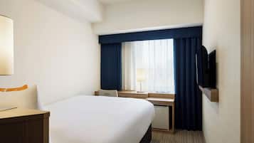 A hotel room with a large bed, a desk, a chair, a TV mounted on the wall, and a window with curtains.