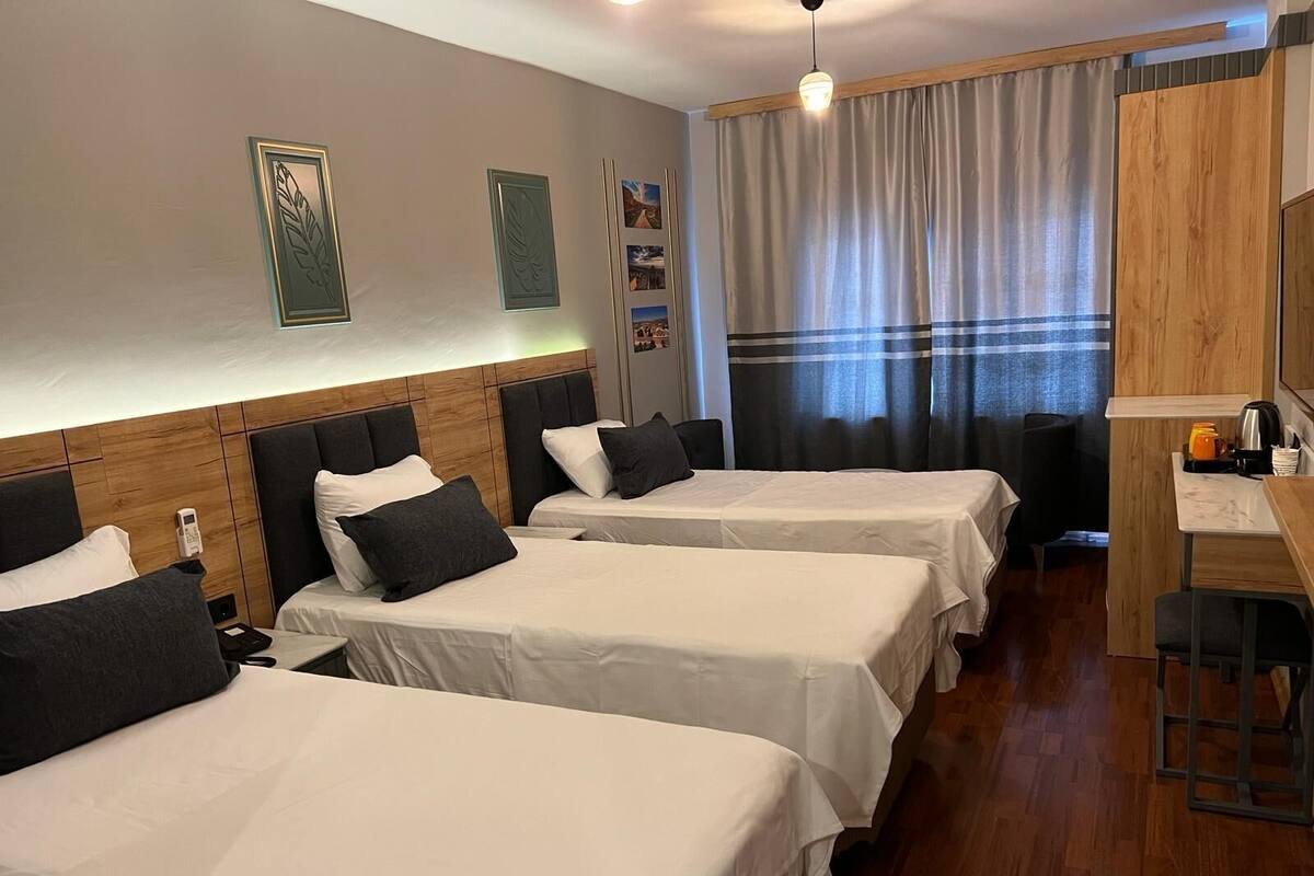 Quarto de hotel com três camas de solteiro, cabeceira de madeira, escrivaninha com cadeira e uma janela grande com cortinas.